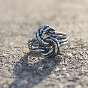 May include: A silver ring with a knot design. The ring has a detailed knot at the centre, with multiple bands forming the ring's base. The ring is set against a textured, grey surface.