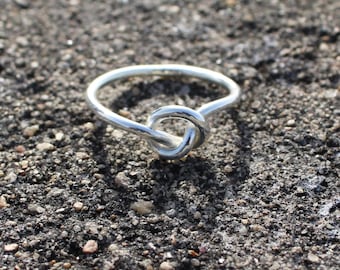 Könnte beinhalten: Ein silberner Ring mit einem Knoten-Design. Der Ring aus glänzendem Metall liegt auf einer strukturierten, dunkelgrauen Oberfläche. Der Knoten ist das zentrale Element des Ringdesigns.