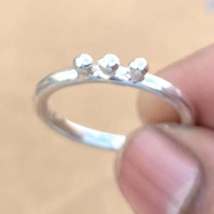 Könnte beinhalten: Ein silberner Ring mit drei kleinen, runden, erhabenen Verzierungen. Der Ring ist glatt und poliert. Der Ring wird zwischen den Fingern gehalten, um sein Design und seine Handwerkskunst zu präsentieren.