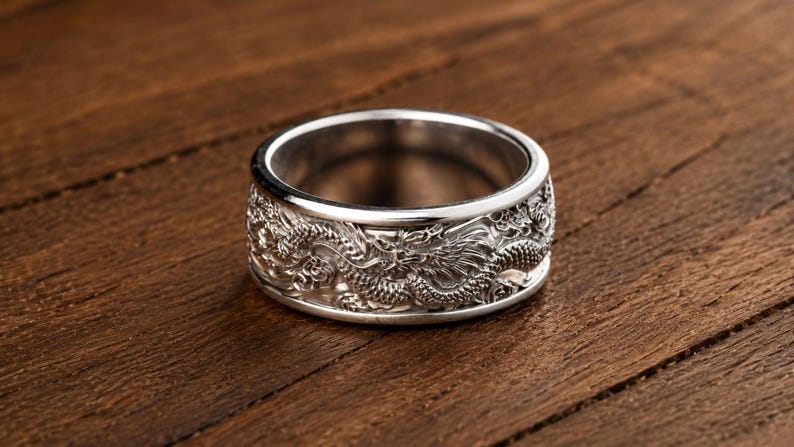 Pu&ograve; includere: Un anello in metallo color argento caratterizzato da un motivo a drago in rilievo dettagliato che avvolge l'intera fascia esterna. La superficie dell'anello ha una finitura lucida con aree incavate che mettono in risalto le squame.