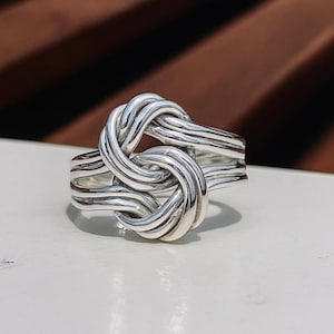 May include: A silver-colored ring with a knot design. The ring features a detailed, interwoven knot in the center, with multiple bands forming the ring's base. The metal has a polished, reflective surface, and the ring is set against a neutral background.