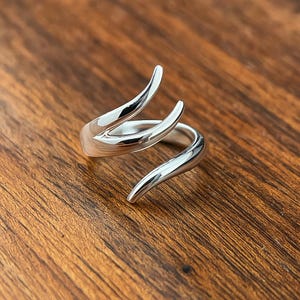 Può includere: Un anello color argento con un design curvo unico. L'anello presenta due elementi prominenti, curvi verso l'alto, che creano uno stile aperto e avvolgente. Il metallo lucido riflette la luce e l'anello è su una superficie di legno.
