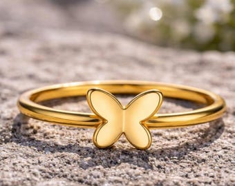 Può includere: Un anello dorato con un design a farfalla. La farfalla è al centro dell'anello e ha una finitura liscia e lucida. L'anello è posto su una superficie grigia strutturata.