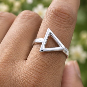 May include: A polished silver ring featuring a prominent open triangle design on the band. The metal surface is smooth and reflective, showcasing a clean geometric shape that sits flat against the finger. The ring has a simple, continuous band.