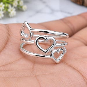 May include: A silver ring featuring three stacked bands, each adorned with a polished open heart cutout. The design consists of three distinct, connected metal loops that create a layered appearance, showcasing a smooth, reflective metallic finish.