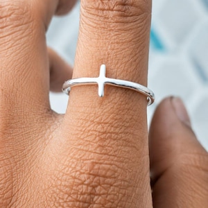 Puede incluir: Un anillo de plata pulida que presenta un diseño de cruz simple centrado en la banda. El anillo tiene un acabado metálico liso y reflectante, con una banda circular delgada que rodea el dedo, mostrando una estética limpia y directa.