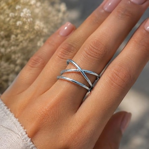 Peut inclure: Une bague argentée au design entrecroisé unique. La bague présente plusieurs bandes qui s'entrecroisent, créant un look ouvert et moderne. La bague est présentée sur une main aux ongles manucurés.