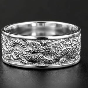 Pu&ograve; includere: Un anello a fascia larga color argento caratterizzato da un intaglio in rilievo dettagliato di un drago che si avvolge attorno all'intera superficie esterna. Il metallo presenta una finitura lucida sui bordi, in contrasto con il design testurizzato.