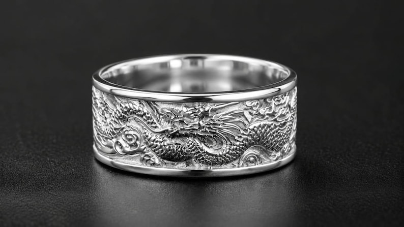 Pu&ograve; includere: Un anello a fascia larga color argento caratterizzato da un intaglio in rilievo dettagliato di un drago che si avvolge attorno all'intera superficie esterna. Il metallo presenta una finitura lucida sui bordi, in contrasto con il design testurizzato.