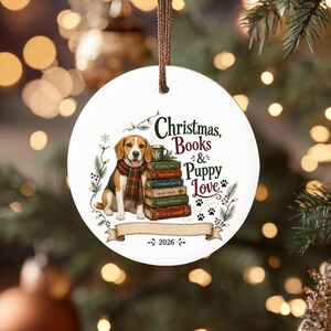 Può includere: Un ornamento rotondo in ceramica bianca appeso a un cordino dorato. Raffigura un beagle con una sciarpa a quadri accanto a una pila di libri e una tazza. Il testo recita Christmas, Books & Puppy Love con impronte di zampe e l'anno 2026.