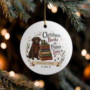 Può includere: Un ornamento rotondo in ceramica bianca con un Labrador marrone che indossa una sciarpa a quadri rossi e verdi accanto a una pila di libri sormontata da una tazza. Il testo recita Christmas, Books & Puppy Love, Your Text Here e 2026 con zampe.