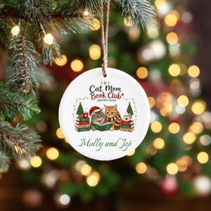 Può includere: Un ornamento rotondo in ceramica bianca appeso a uno spago marrone. Il design mostra due gatti, uno con un cappello da Babbo Natale e uno con una sciarpa, che leggono libri. Il testo recita Cat Mom Book Club Christmas Edition e Molly and Jep.