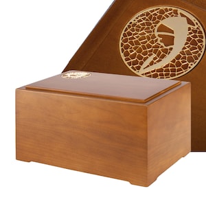 Könnte beinhalten: Eine hellbraune Urne aus Holz mit rechteckiger Form. Der Deckel hat ein kleines, kreisförmiges goldenes Emblem. Hinter der Urne befindet sich eine dekorative Tafel mit einem goldenen Emblem und einem gemusterten Hintergrund.