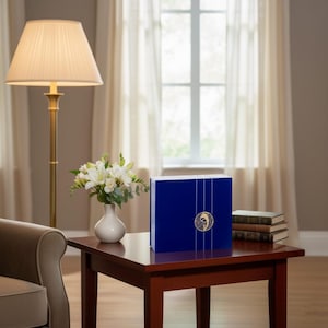 Könnte beinhalten: Eine marineblaue Box mit goldenen Akzenten, darunter ein kreisförmiges Emblem und vertikale Linien. Die Box steht auf einem dunklen Holztisch, neben einer Vase mit weißen Blumen und einem Stapel Bücher. Eine Stehlampe und ein Fenster sind im Hintergrund.