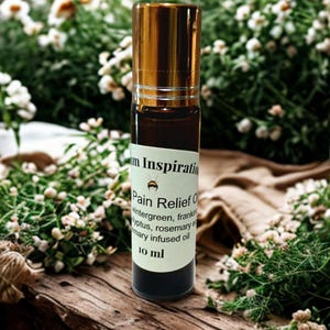 Puede incluir: Un frasco roll-on de vidrio marrón con tapa dorada, etiquetado como "Pain Relief Oil". El frasco se encuentra sobre un fondo de flores blancas, vegetación y una superficie de madera. La etiqueta enumera los ingredientes y el volumen es de 10 ml.