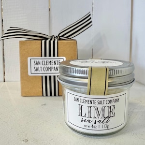 Puede incluir: Un tarro de sal marina de lima con tapa plateada y una etiqueta que dice "San Clemente Salt Company" y "Lime Sea Salt, Net Wt 4oz | 113g." En el fondo, una pequeña caja de regalo con una cinta a rayas blancas y negras.