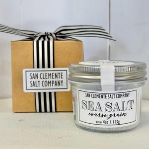 May include: A jar of coarse grain sea salt with a silver lid and a label that reads "San Clemente Salt Company Sea Salt". A small gift box with a black and white striped ribbon is behind the jar.