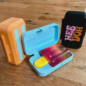 May include: Three rectangular plastic cases shaped like popsicles. One is orange with light blue accents, one is light blue and open containing a red and yellow popsicle-shaped object, and one is black with the pink and orange text NEE DOH.