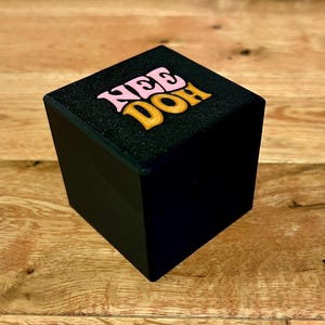 Könnte beinhalten: Eine schwarze quadratische Box mit dem Wort "NEE DOH" in rosa und orangefarbenen Buchstaben auf der Oberseite. Die Box steht auf einer Holzoberfläche.