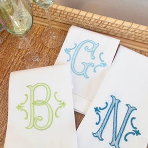 May include: Three white rectangular cloth napkins featuring embroidered monograms in different colors. One napkin displays a light green letter B, another a light blue letter G, and the third a dark blue letter N, each with decorative scrollwork.