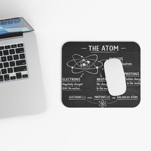Op de afbeelding: Zwart rechthoekig muismatje met een witte muis en een diagram van een atoom. Het diagram bevat labels voor elektronen, neutronen en protonen. De tekst "THE ATOM" staat bovenaan.