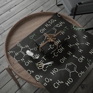 May include: A wrapped present with a brown ribbon sits on black wrapping paper printed with white chemical formulas. A roll of tape and gold-handled scissors are also on the paper, which is on a wooden table.