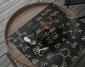 May include: A wrapped gift with a brown ribbon sits on black wrapping paper printed with white chemical formulas. A roll of tape and gold-handled scissors are also on the paper, which is on a wooden table.