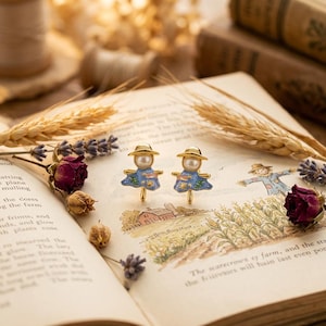 May include: A pair of gold-toned stud earrings shaped like scarecrows, each with a pearl head and a blue dress with floral details. The earrings are set on an open book with illustrations and dried flowers.