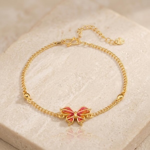 May include: Gold-toned bracelet featuring a butterfly charm with red and orange enamel detailing. The bracelet has a delicate chain design with a lobster clasp closure and adjustable length. Two small gold beads are placed on the chain.