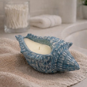 Könnte beinhalten: Ein blaues Muschel-Kerzenhalter mit einer cremefarbenen Kerze. Der Kerzenhalter steht auf einem beigen Handtuch in einem Badezimmer. Das Muscheldesign hat strukturierte Details. Die Kerze ist aus Wachs.