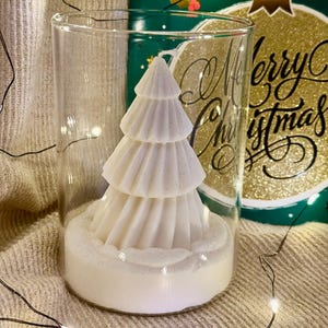 Könnte beinhalten: Eine weiße, mehrschichtige Weihnachtsbaumkerze mit Glitzerakzenten befindet sich in einem transparenten Glaszylinder. Die Kerze steht auf einem weißen Sockel, im Hintergrund eine Weihnachtskarte mit der Aufschrift "Merry Christmas."