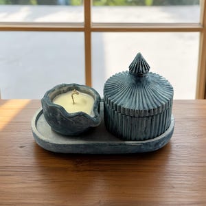 Puede incluir: Un juego de velas azul grisáceo sobre una bandeja a juego. El juego incluye un pequeño portavelas festoneado con una vela encendida y un tarro con tapa con un diseño texturizado y acanalado. Los artículos están sobre una superficie de madera.