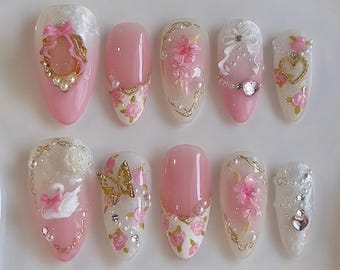 May include: A set of ten almond-shaped press-on nails featuring a pink and white color palette. The designs include 3D elements such as gold frames, white bows, pink floral accents, a white swan, a gold butterfly, small pearls, and clear rhinestones.