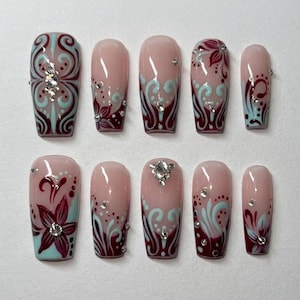 Peut inclure: Un ensemble de dix faux ongles avec une base nude, ornés de motifs tourbillonnants bordeaux et bleu clair, de fleurs et de strass transparents. Les ongles présentent une finition brillante et une forme carrée effilée.