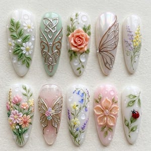 May include: A collection of ten almond-shaped, decorated fingernails. Each nail features a unique design, including floral arrangements, butterflies, and Celtic knotwork, with pearl and rhinestone accents. The color palette includes white, pink, green, and silver.