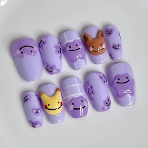 May include: A set of ten lavender-coloured artificial nails. Each nail features a different design, including cartoon characters and smiley faces. Some nails have 3D elements, and others have floral patterns.