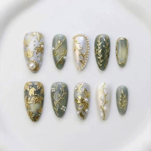 May include: A collection of ten artificial nails in various shapes and sizes. Each nail features a unique design with gold leaf accents, pearls, and intricate patterns. The color palette includes shades of green, white, and gold, creating an elegant and sophisticated look.