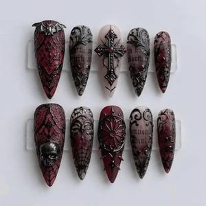 May include: A set of ten stiletto-shaped press-on nails featuring a dark red and black color palette. The designs include raised metallic embellishments such as a bat, a skull, a cross, and intricate filigree patterns over a textured lace base.