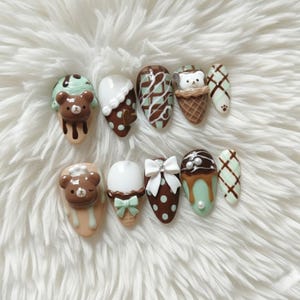 May include: A set of ten artificial nails with a dessert theme. Designs include ice cream cones, teddy bears, and decorative patterns in brown, white, and mint green. The nails are set against a fluffy white background.