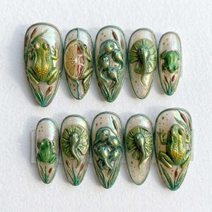 Può includere: Set di dieci unghie finte a tema natura. Ogni unghia presenta un design diverso, tra cui rane, girini e ninfee, nelle tonalità del verde e dell'oro. Le unghie hanno una base perlescente e sono decorate con dettagliata arte in rilievo.