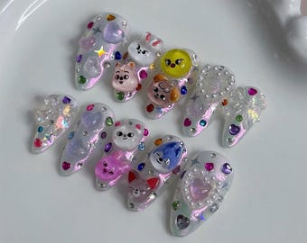 May include: A set of artificial nails with a pearlescent finish. Each nail features a different cartoon character face, along with iridescent gems and star-shaped accents. The nail shapes are oval and vary in size.