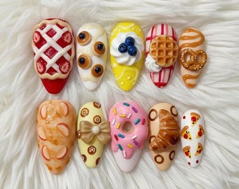 Peut inclure: Dix faux ongles décorés de motifs alimentaires miniatures. On retrouve une tarte aux fraises, des crêpes, un cupcake aux myrtilles, une gaufre, un bretzel, une pêche, un donut, un croissant et une pizza.