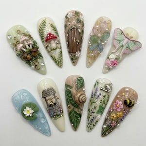 Op de afbeelding: Een set van tien lange, amandelvormige kunstnagels, elk uniek versierd met miniatuurontwerpen. De ontwerpen omvatten paddenstoelen, een sprookjesdeur, een slak, een put, een mot en bloemen, allemaal in een pastelkleurenpalet.