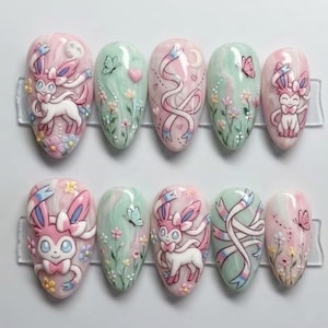 May include: A set of ten almond-shaped press-on nails featuring a pastel color palette of pink, white, and mint green. The designs include 3D-style illustrations of the character Sylveon, swirling ribbons, small butterflies, and delicate floral motifs.