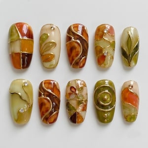 May include: A set of ten press-on nails featuring a color palette of olive green, amber, and cream. The designs include gold metallic line work, swirling patterns, floral motifs, and small embedded clear rhinestones on various nail surfaces.
