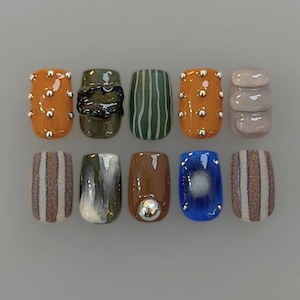 Peut inclure: Un ensemble de dix faux ongles présentant des motifs variés, notamment orange avec des clous argentés, vert foncé avec une texture noire, vert à rayures blanches, beige avec des lignes en relief, marron à rayures pailletées et bleu avec une sphère argentée.