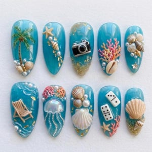 May include: A collection of ten turquoise artificial nails, each almond-shaped and decorated with a summer theme. Designs include seashells, starfish, coral, a camera, a palm tree, and a beach chair, with pearl embellishments.