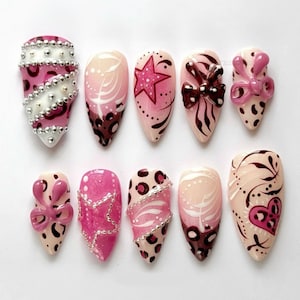 May include: A set of ten almond-shaped press-on nails featuring a variety of designs in shades of pink, white, and brown. The nails are decorated with leopard print patterns, raised pink bows, silver metallic beads, glittery stars, and heart motifs.
