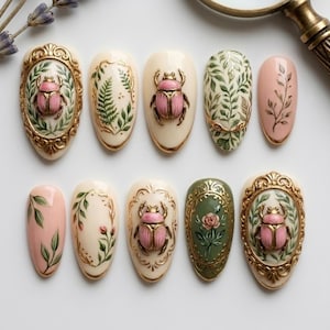 May include: A collection of ten oval-shaped nail art designs. The designs feature a variety of elements, including pink beetles, green leaves, and floral patterns, all set against a cream-colored base with gold accents. The nails are arranged on a white surface.