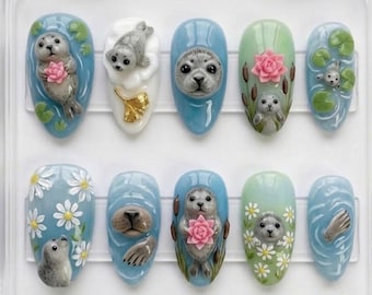Puede incluir: Diez uñas artificiales con diseños detallados de focas en 3D. Cada uña presenta una escena diferente, incluyendo focas con flores, en el agua y entre plantas. Las uñas están pintadas en tonos de azul, blanco y verde.